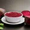 A bowl of beetroot malt powder with fresh beetroots and leaves on a gray background.